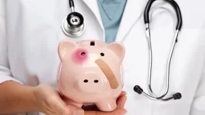 The image focuses on a piggy-bank in the hand of a doctor. The piggy bank has a black eye and a bandage indicating the financial decreases for providers due to Medicare price cuts and inflation.